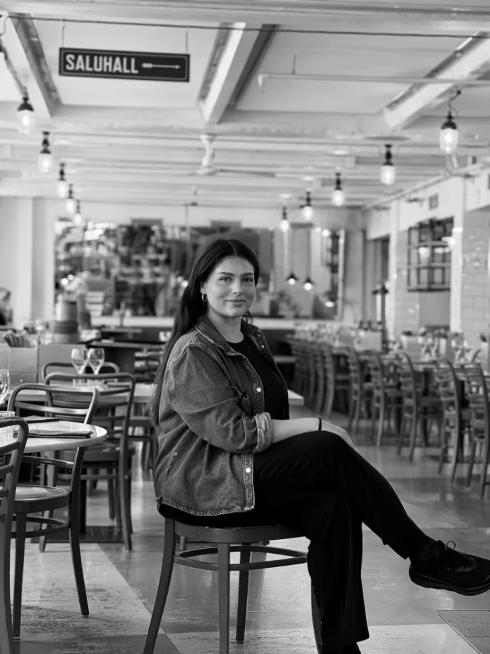 Medarbetare på B.A.R. i mörk jacka och mörkt hår, sittande i matsalen under skylten Saluhall